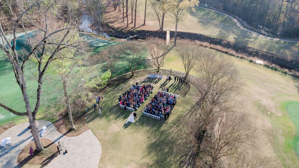 Aerial drone view of an outdoor wedding ceremony with guests seated in rows on a green lawn surrounded by trees.