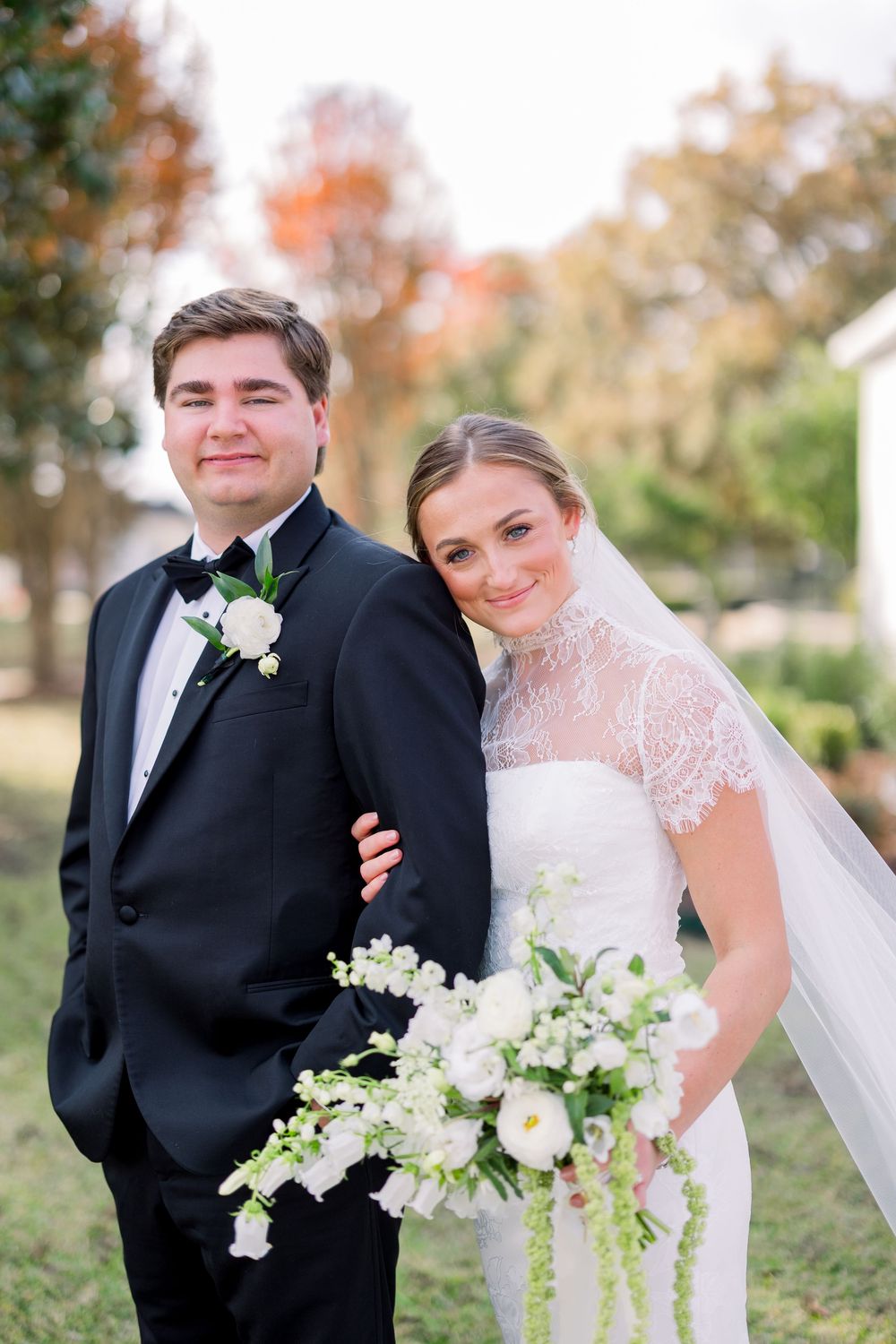 A bride and groom pose together outdoors during fall with white floral bouquet.