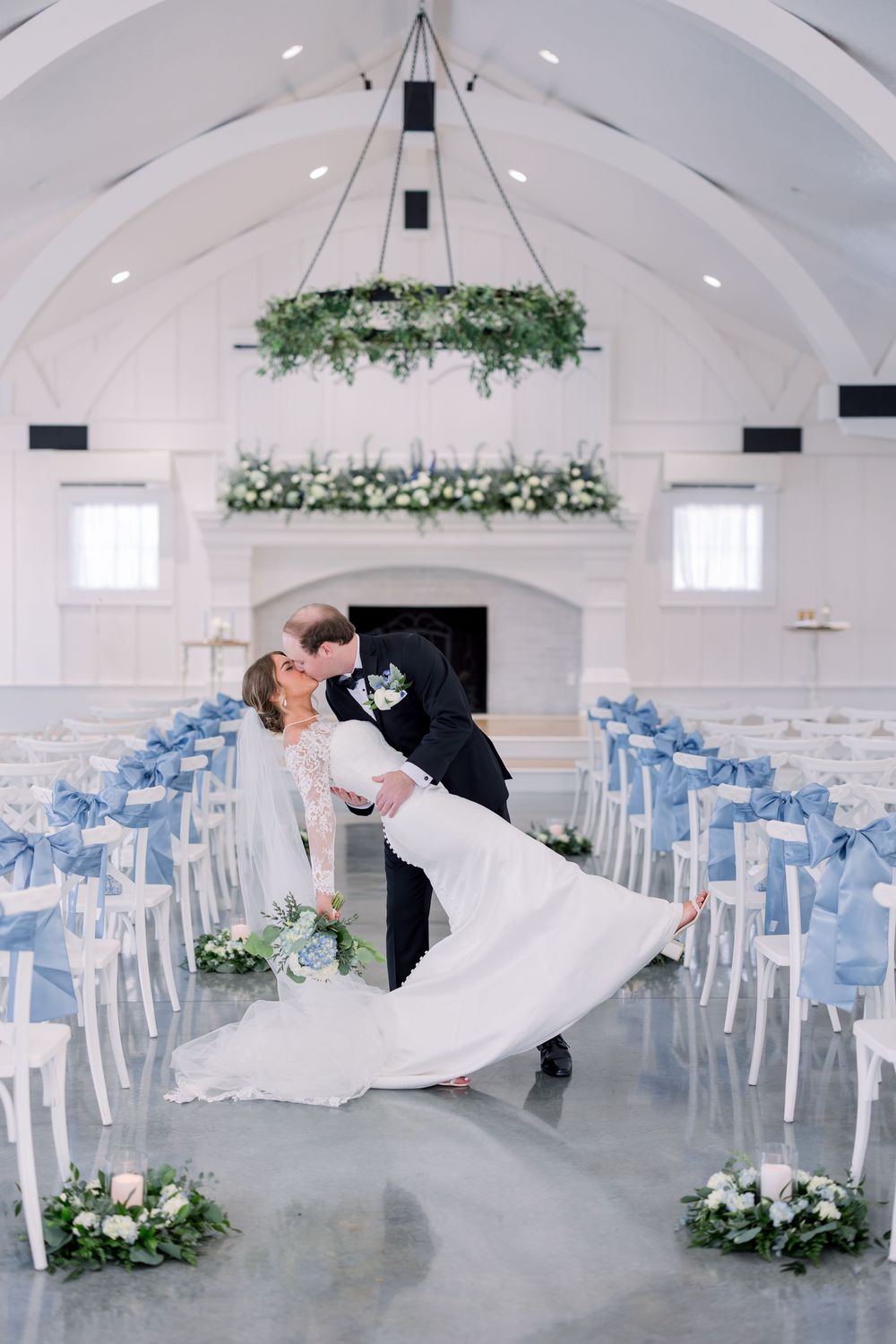 A romantic dip kiss in a white chapel with blue chairs and hanging greenery decorations.