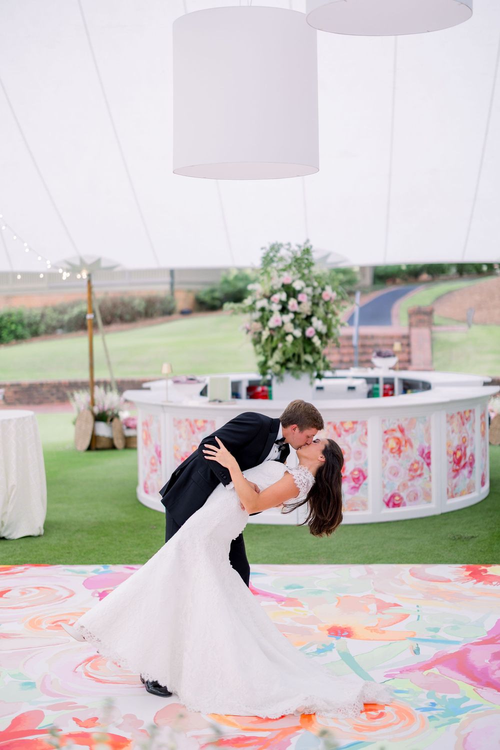 Romantic wedding couple shares a dip kiss on a floral-patterned dance floor under white tent canopies.