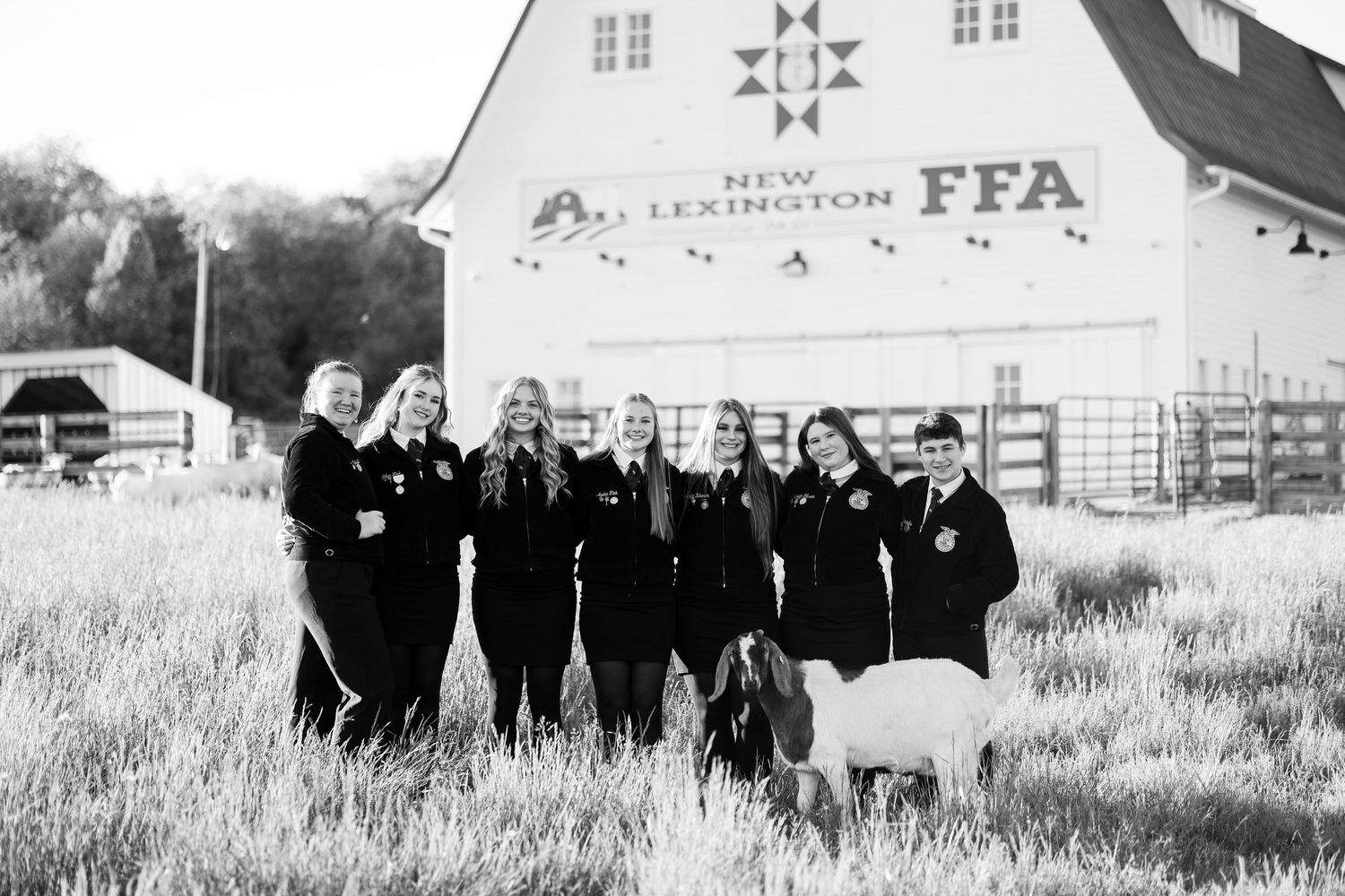 Black and white photo of FFA students in jackets posing with a goat in front of New Lexington FFA barn.