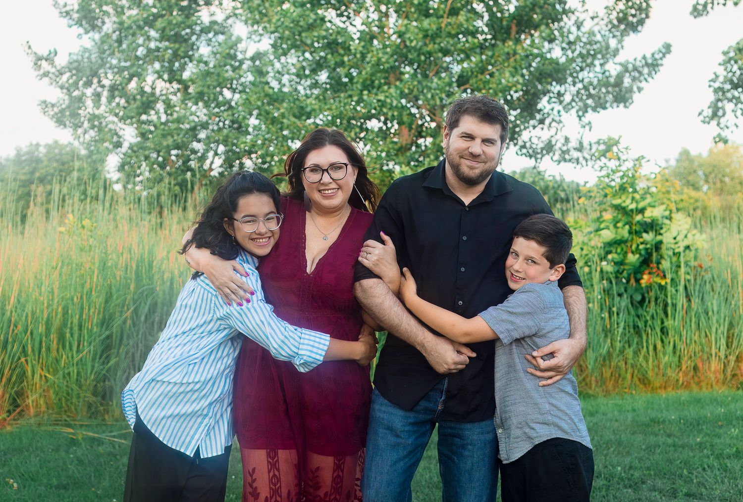 A family of four stands outside, smiling and embracing. They are in front of tall grass and trees.