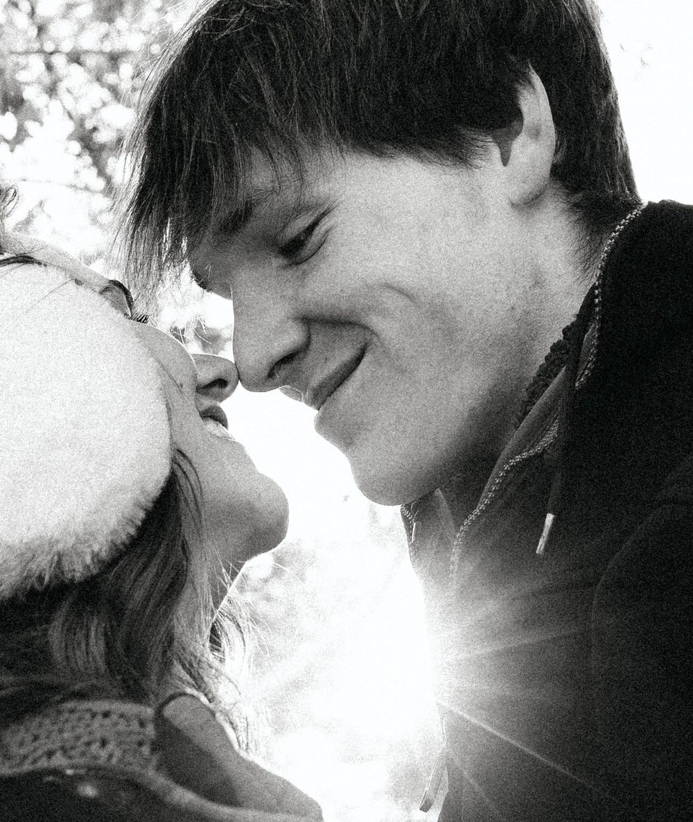 Black and white photo of a smiling couple about to kiss outdoors in winter, with sunlight flaring between them.