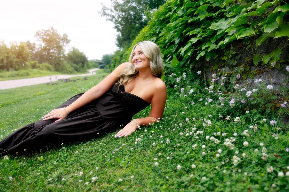 Young blonde woman in black dress lying on green grass with white flowers against ivy-covered stone wall outdoors.