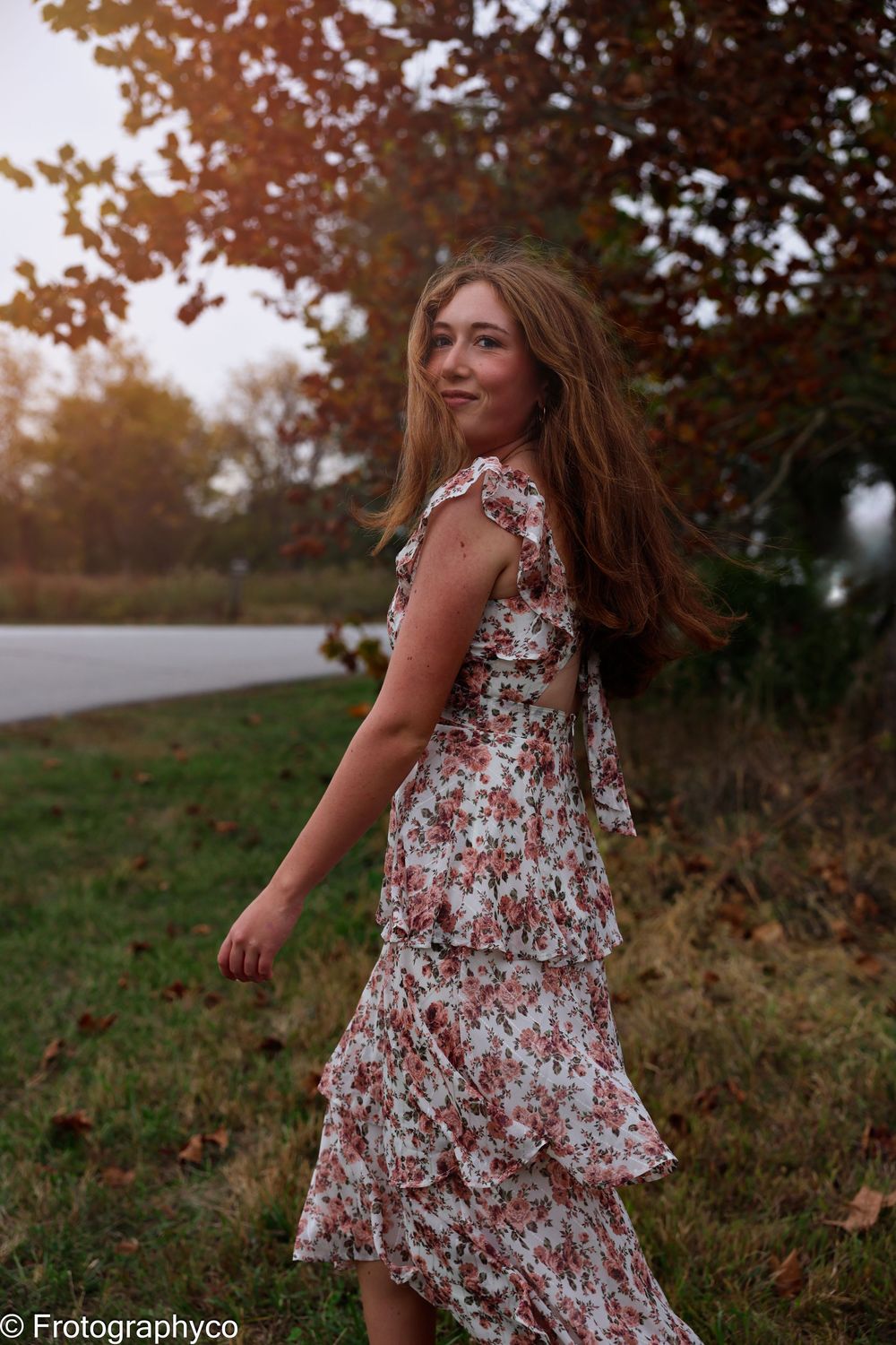 Young woman in floral ruffle dress poses outdoors among autumn trees with fallen leaves on ground.