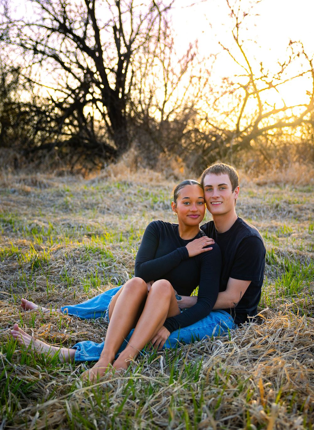 A young couple sitting together in a golden field at sunset, both wearing black tops and jeans, smiling warmly.