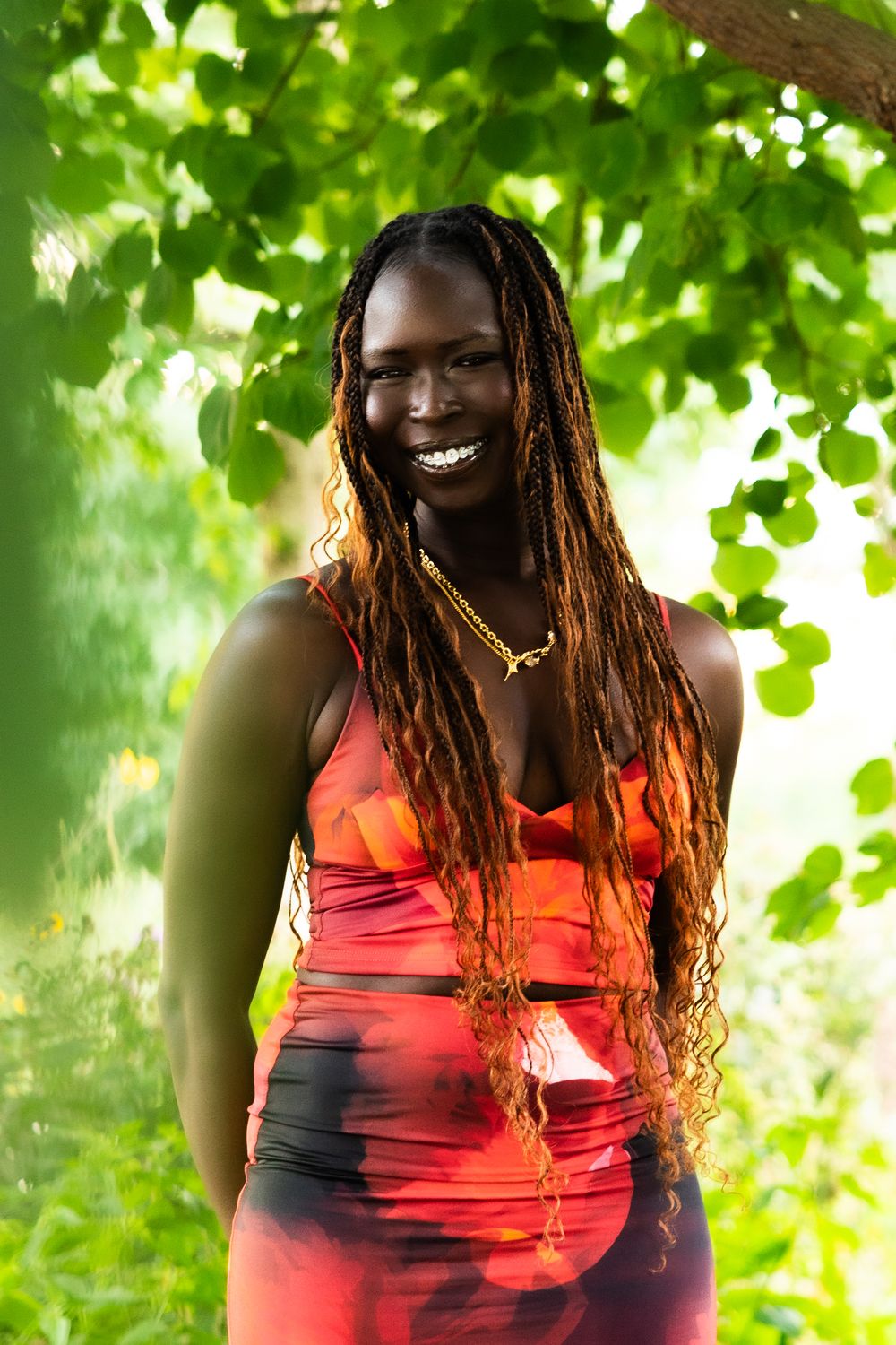 A person in a red dress stands under green foliage with sunlight filtering through the leaves.