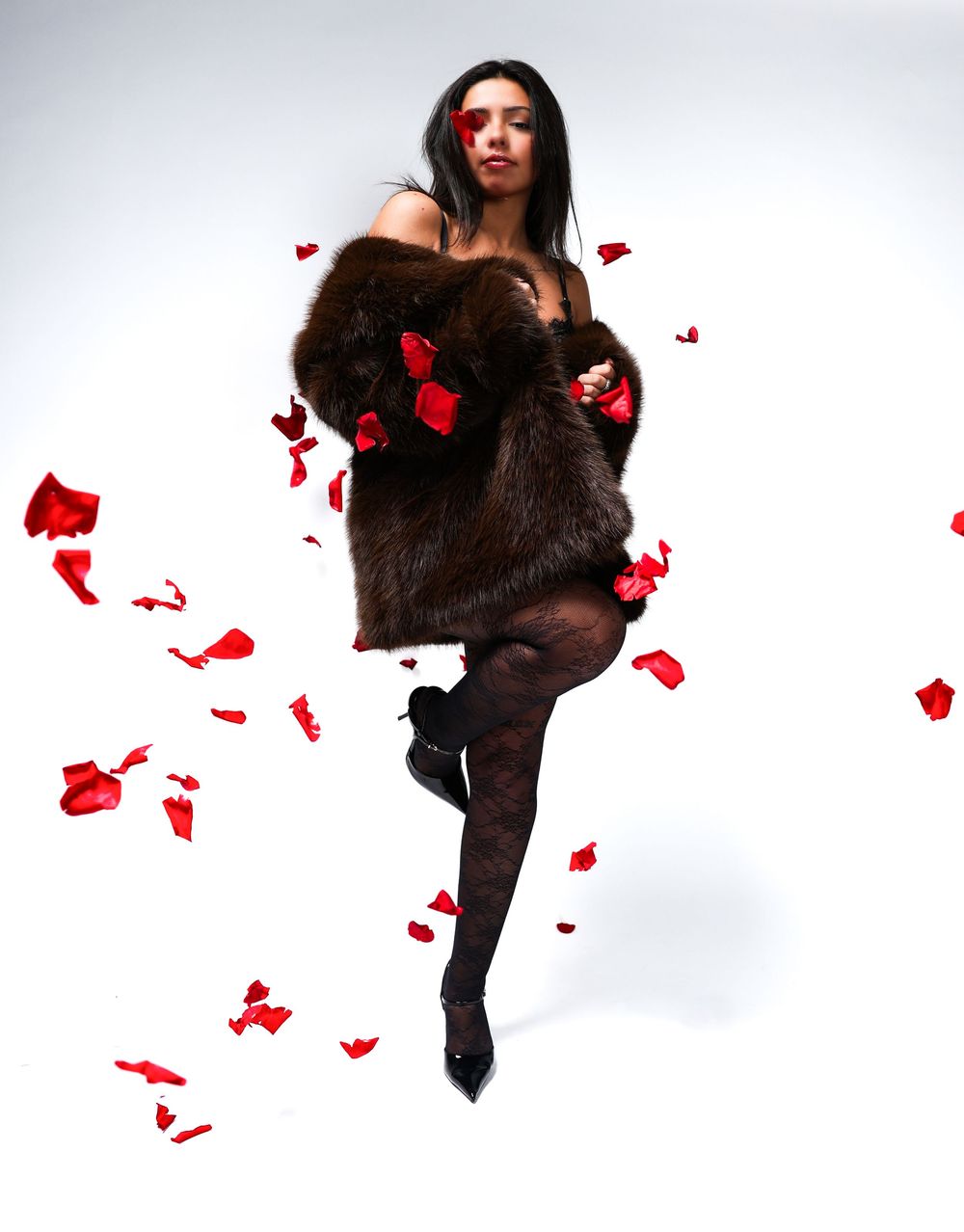 Woman in brown fur coat with red rose petals falling around her, wearing black tights and heels on white background.