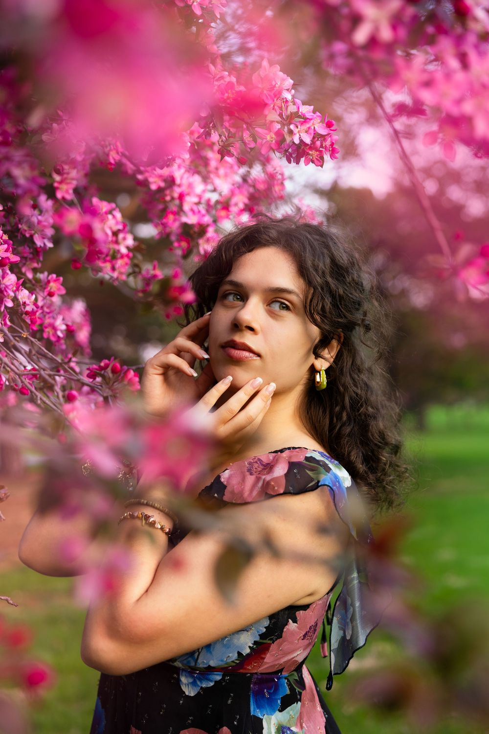 A person poses thoughtfully among blooming pink cherry blossoms wearing a floral dress.