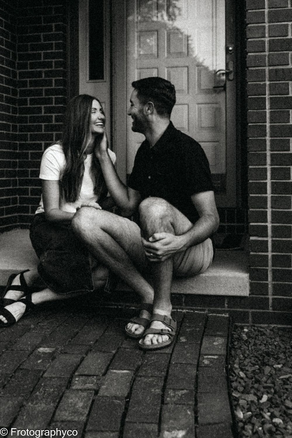 Black and white photo of a smiling couple sitting on brick steps in front of a door, sharing a joyful moment.