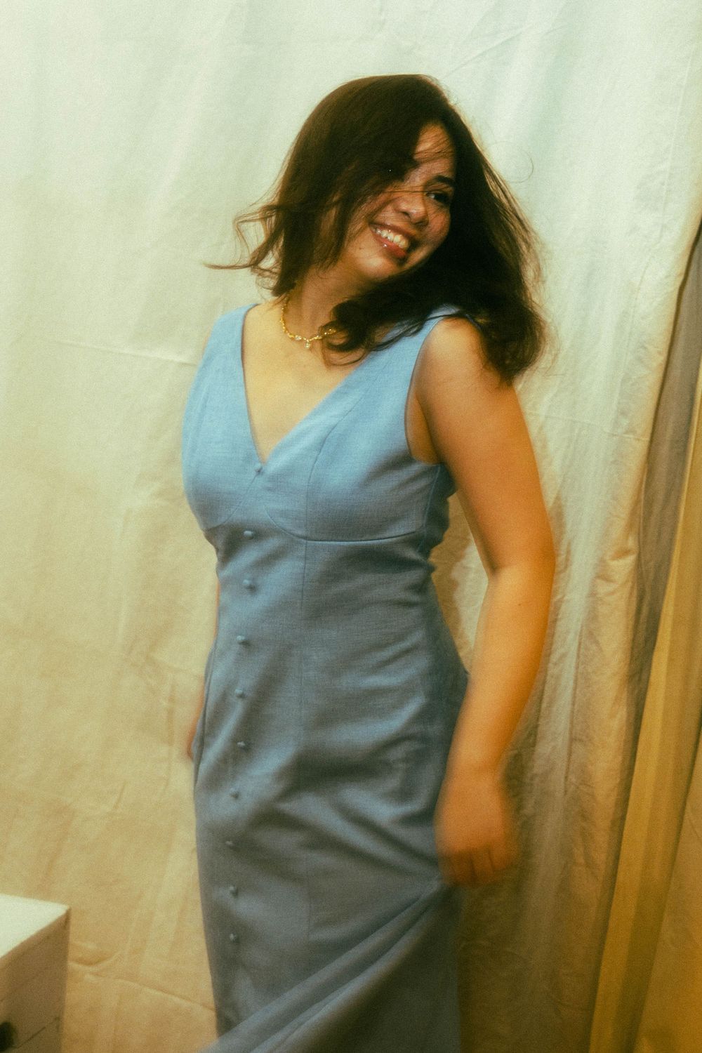 A smiling woman with dark hair poses in a sleeveless blue button-front midi dress against a white curtain backdrop.