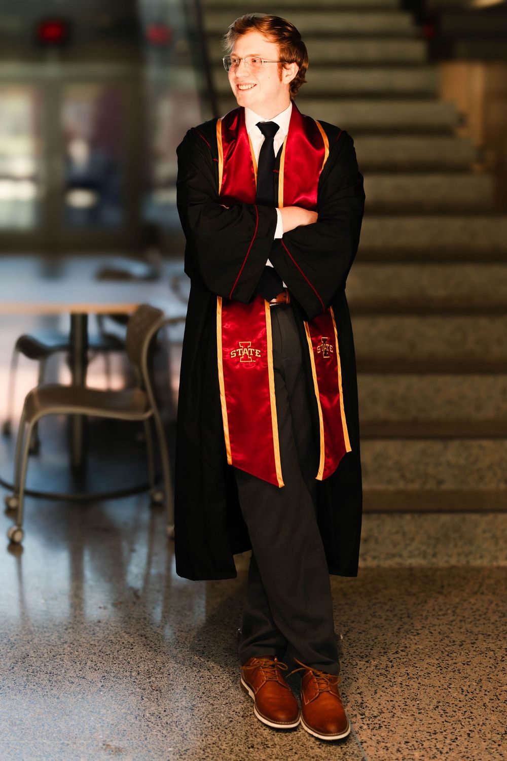 Young man in Iowa State graduation regalia with red stole, black robe, standing with arms crossed, smiling indoors.