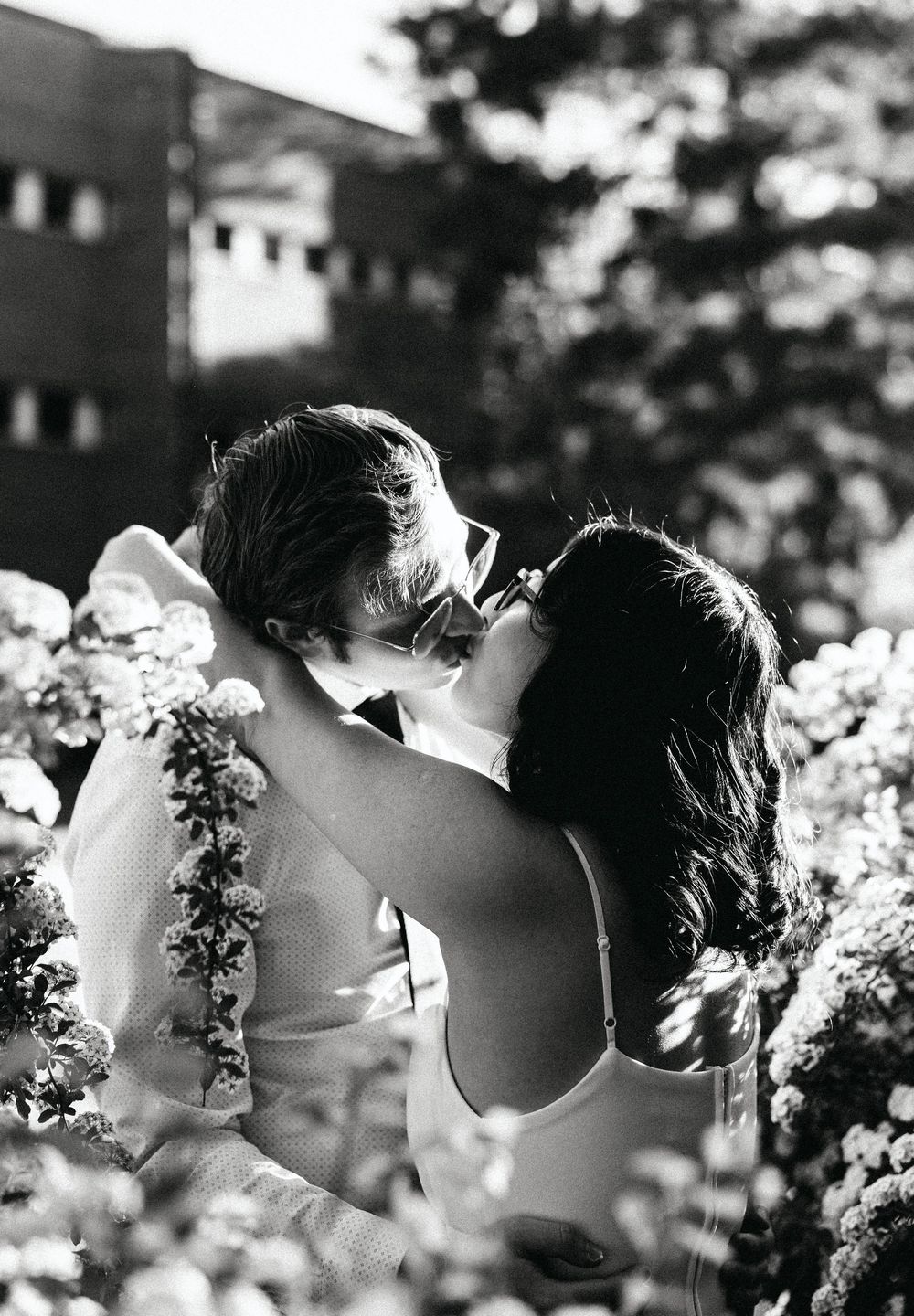 Black and white photo of a couple sharing a romantic kiss surrounded by blooming flowers outdoors.