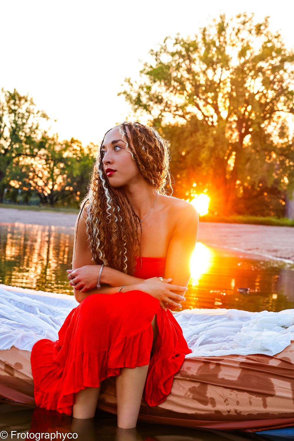 Young woman in red dress sitting by a sunlit lake at sunset with curly hair and trees in the background.