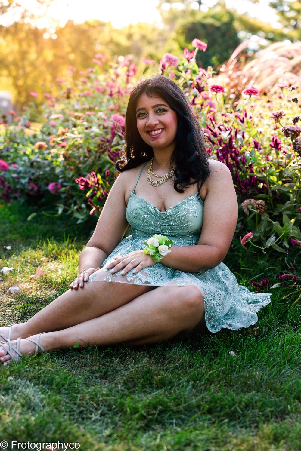 Young woman in mint green dress sitting on grass surrounded by colorful garden flowers, wearing wrist corsage.