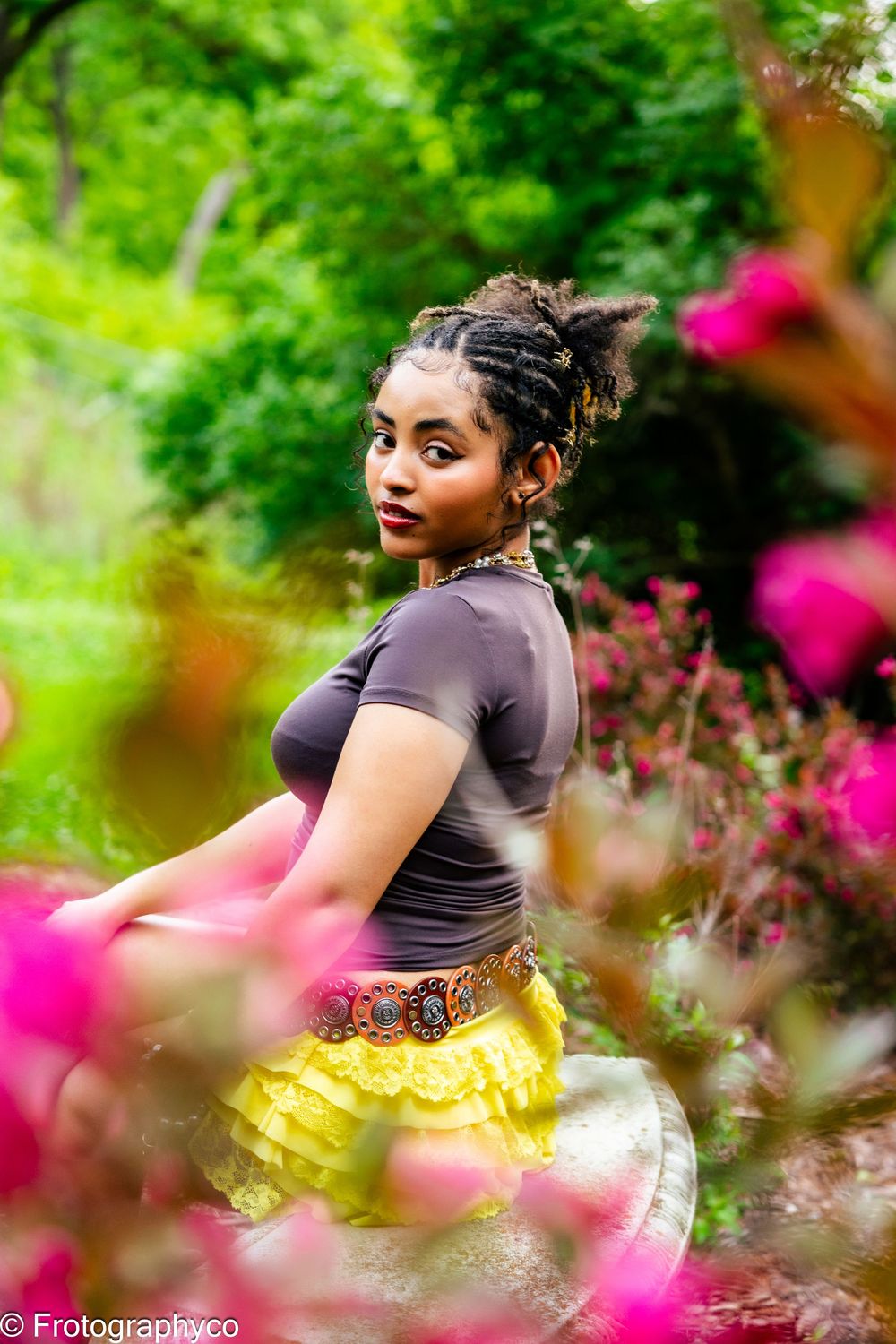 Outdoor portrait through pink flowers showing a person in black top and yellow skirt against lush green garden background.