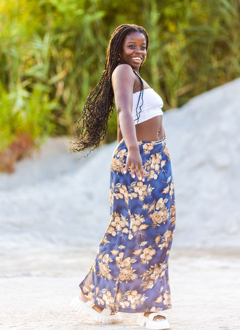 A woman in a white crop top and floral maxi skirt poses outdoors with a bright smile.