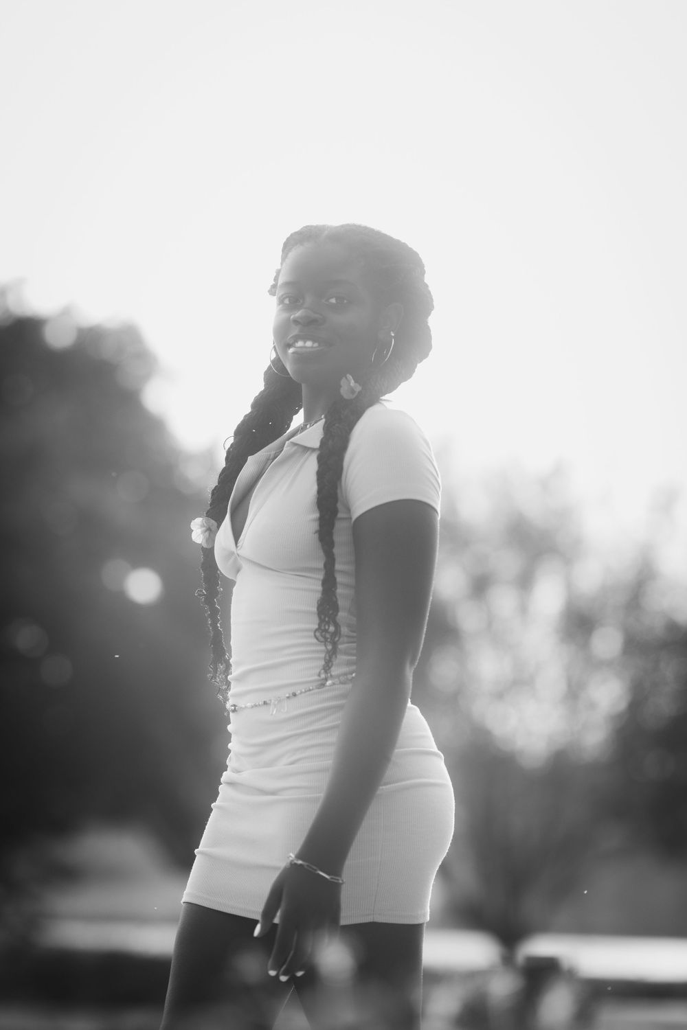 Black and white portrait of a young woman in a white dress with braided hair posing outdoors in natural light.