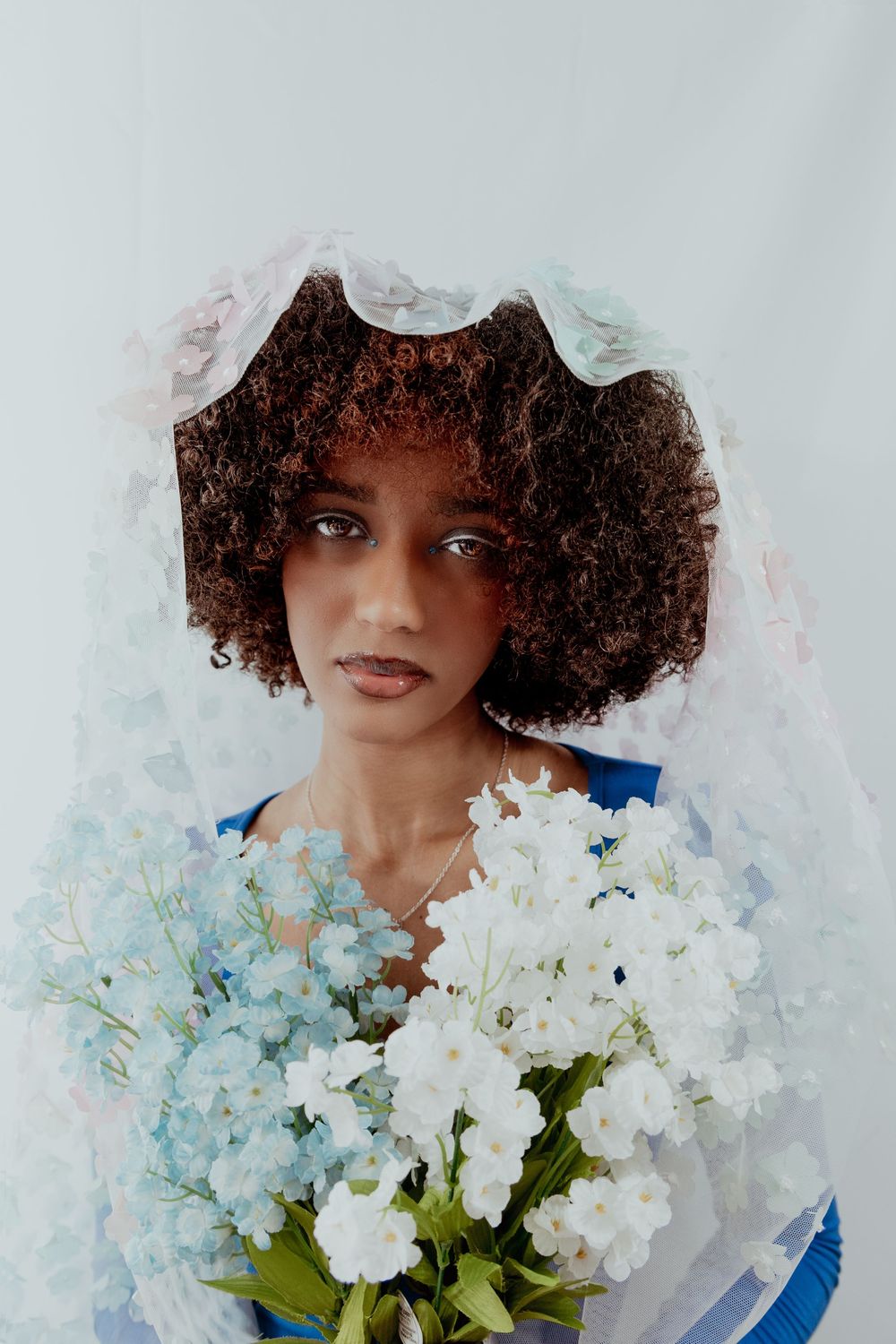 A person with curly hair poses with white flowers against a light backdrop wearing a blue top.