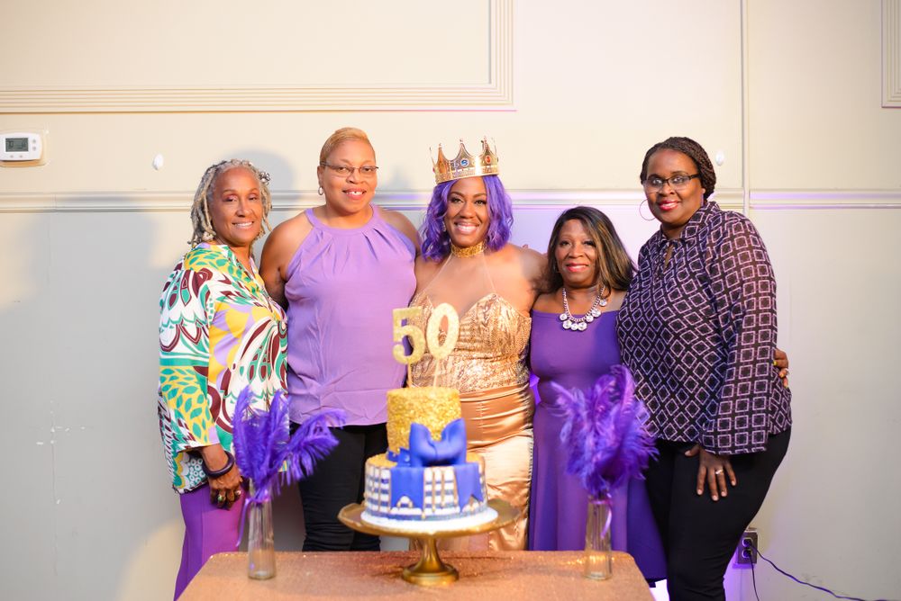 Party guests gathered around a celebratory cake at an event with purple decor.