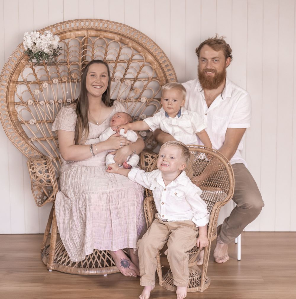 Family portrait of parents with two toddlers and newborn baby seated in a rattan peacock chair against white backdrop.