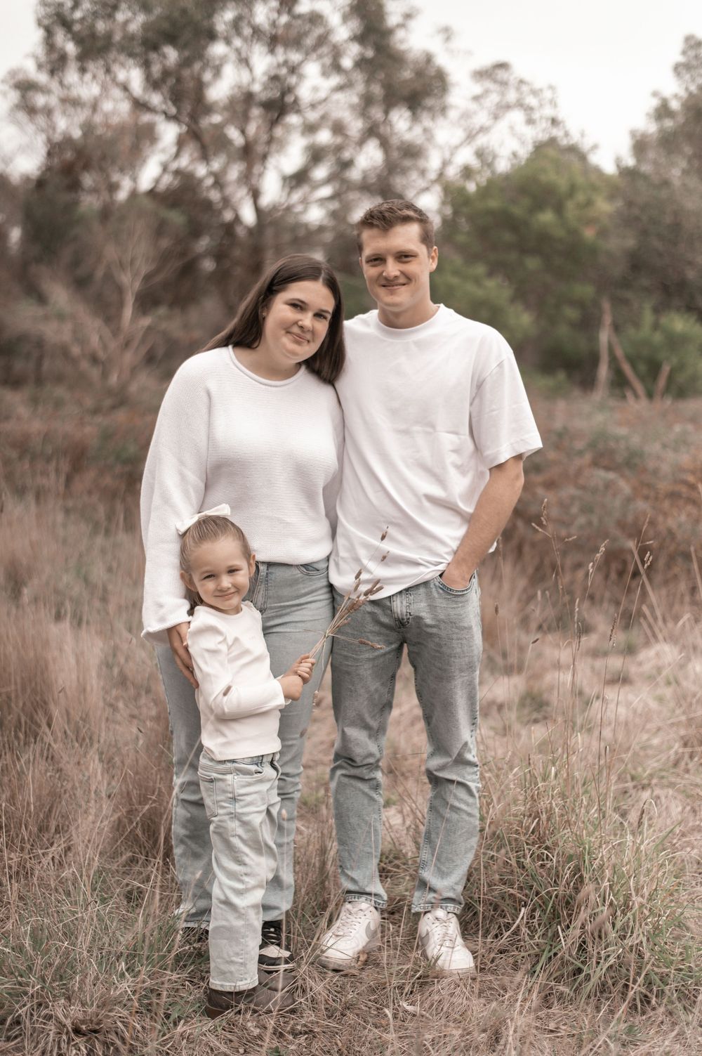 Family of three in white tops and jeans posing outdoors in a natural bush setting, smiling warmly at the camera.