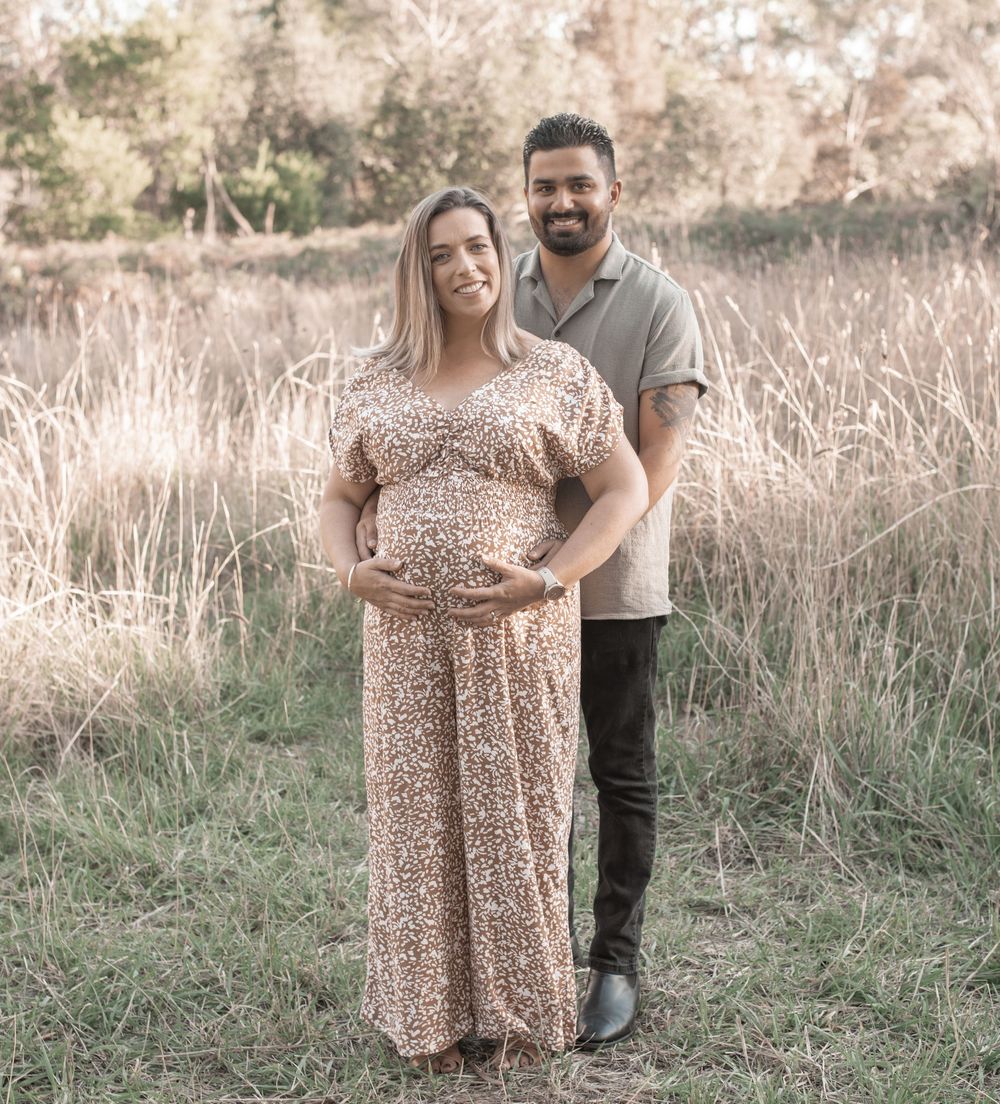 Pregnant woman in floral dress with partner standing in golden grass field during maternity photoshoot outdoors.