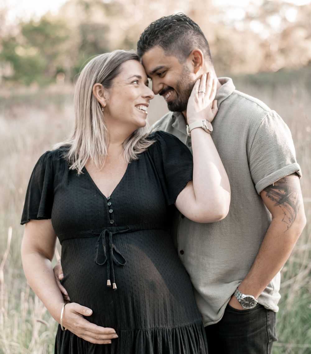 A smiling pregnant woman in a black dress with her partner in a field during a maternity photo shoot.