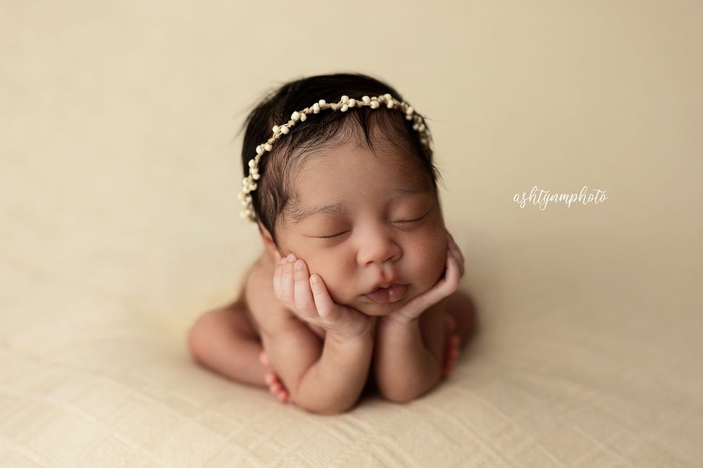 Sleeping newborn baby girl wearing a delicate floral headband, posed with hands under chin on a cream backdrop.