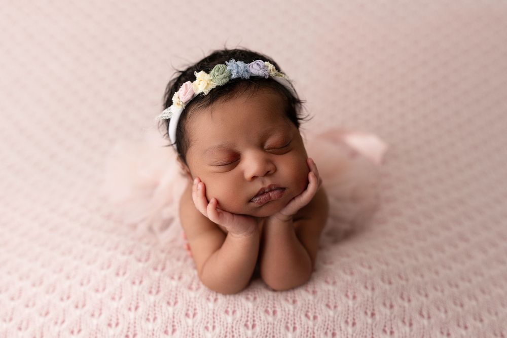 Newborn baby in Colorado Springs wearing white floral headband sleeps peacefully on pink textured blanket with hands under chin.