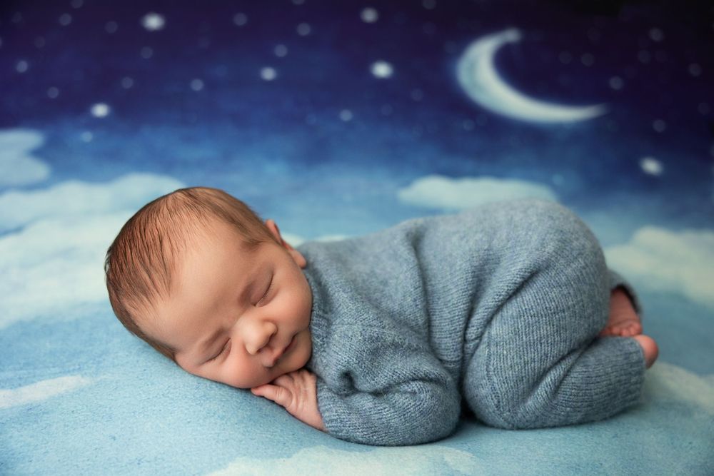 Newborn baby sleeping peacefully in blue outfit on cloud backdrop with crescent moon and stars.