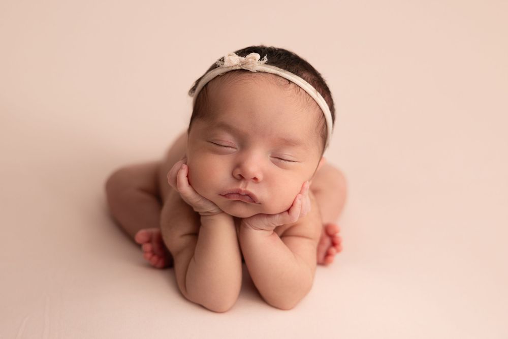 Sleeping newborn baby in Colorado Springs wearing delicate headband poses with hands under chin on pink background.