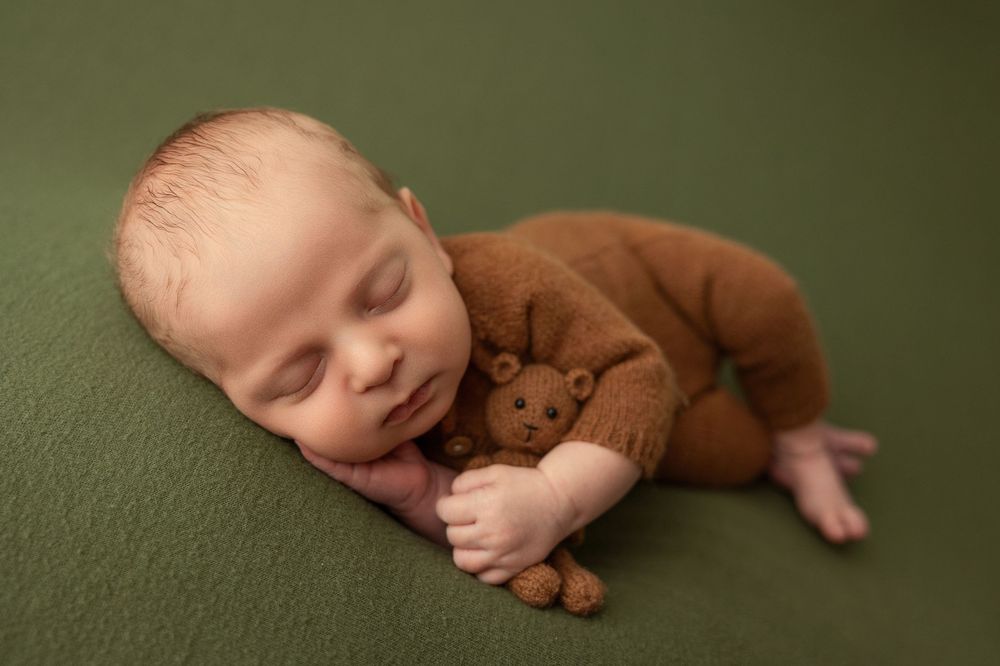 Sleeping baby curled up on soft green blanket with teddy bear in peaceful slumber.