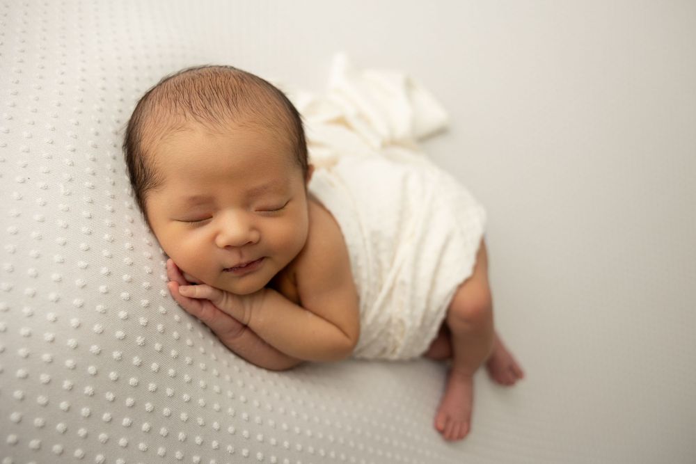 A sleeping newborn baby wrapped in white swaddle cloth lies peacefully against a textured background.