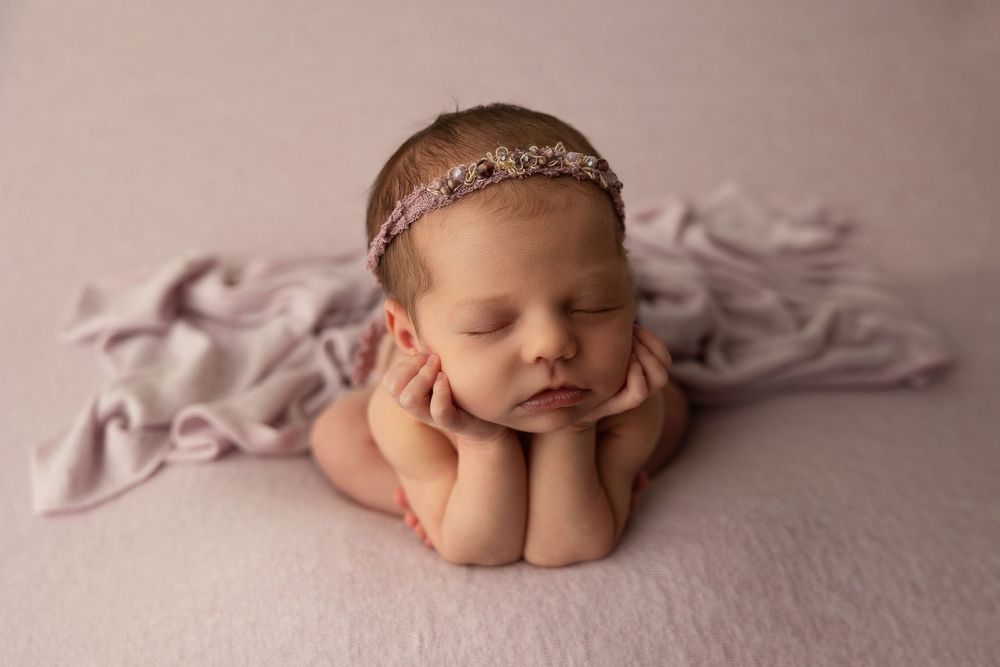 Sleeping newborn baby girl wearing a delicate floral headband, posed with hands under chin on a soft lavender backdrop.