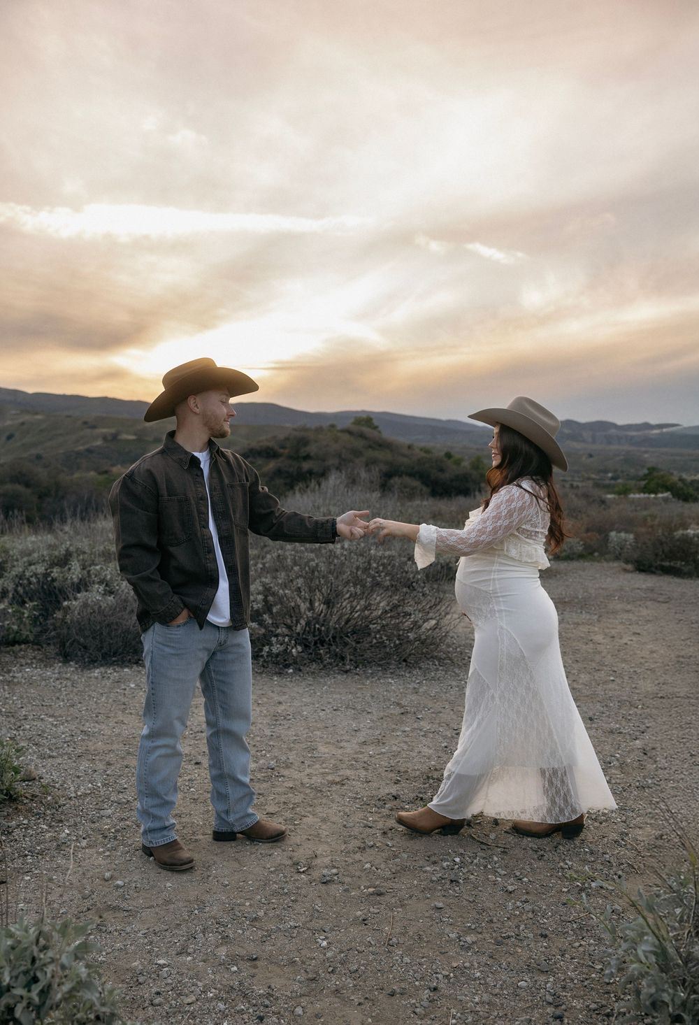 A couple in western attire dances together in a desert landscape at sunset, creating a romantic silhouette against the sky.