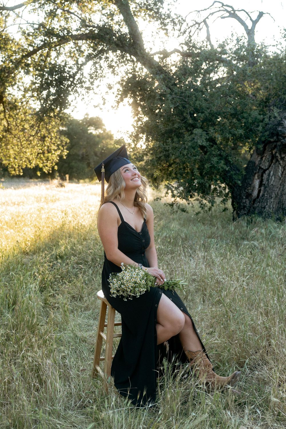 Smiling graduate in black dress and cap sits on chair in sunlit meadow holding white wildflowers near oak tree.