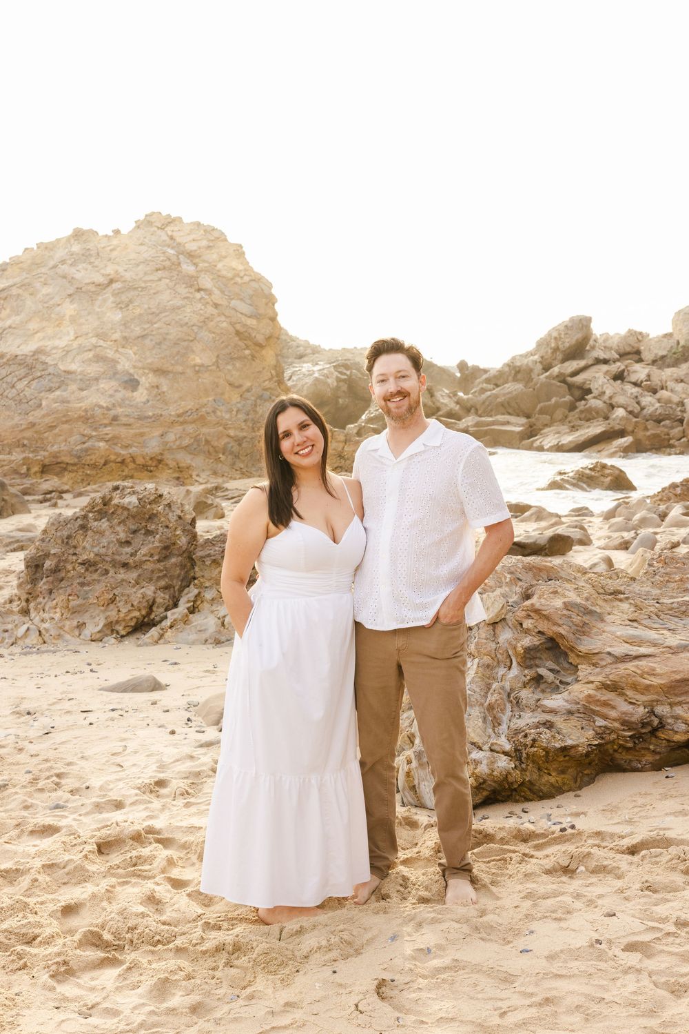 Couple posing on sandy beach with rocky backdrop, woman in white dress and man in white shirt and tan pants.