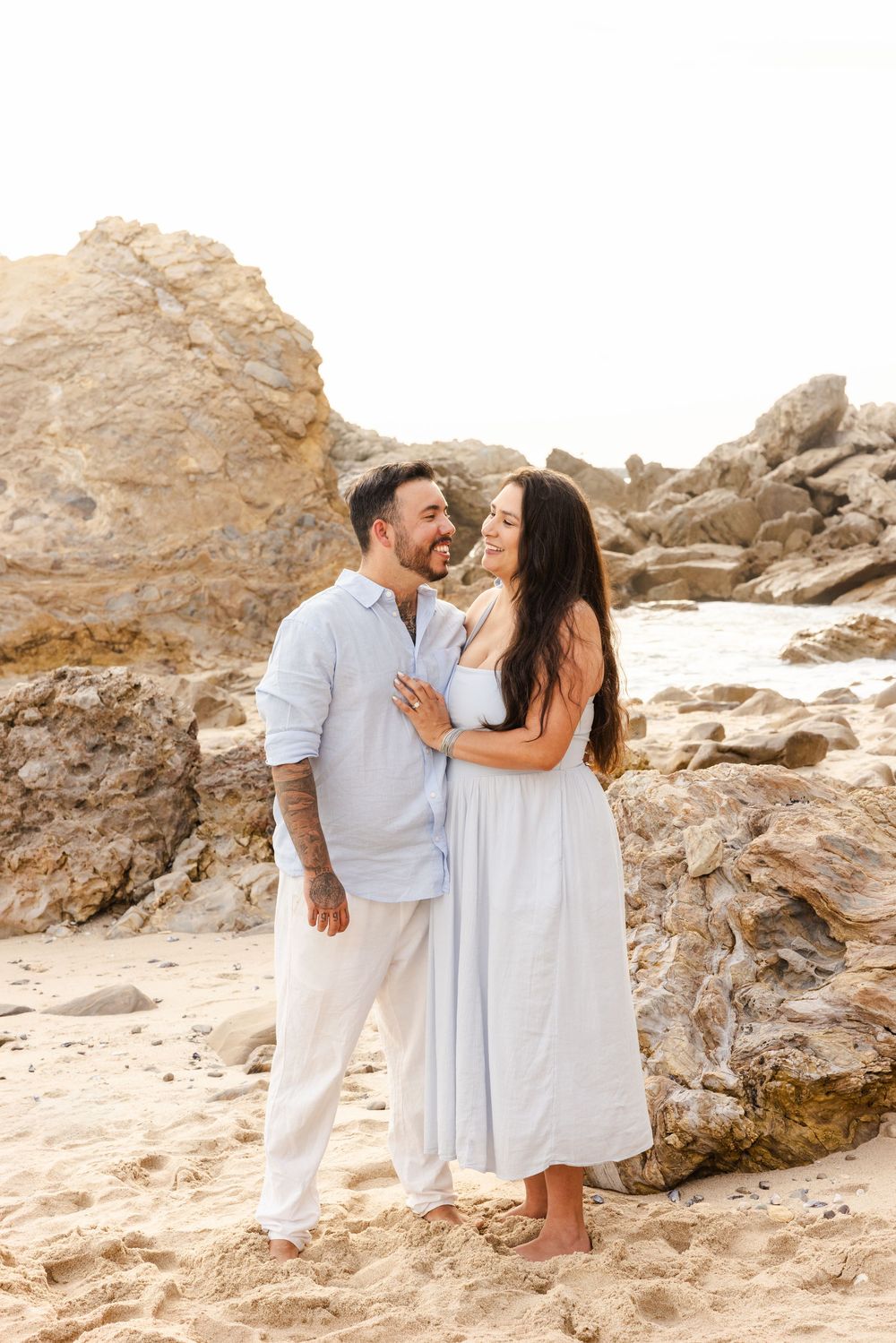 A couple embraces on a sandy beach with rocky formations, wearing white and light blue outfits, smiling at each other.