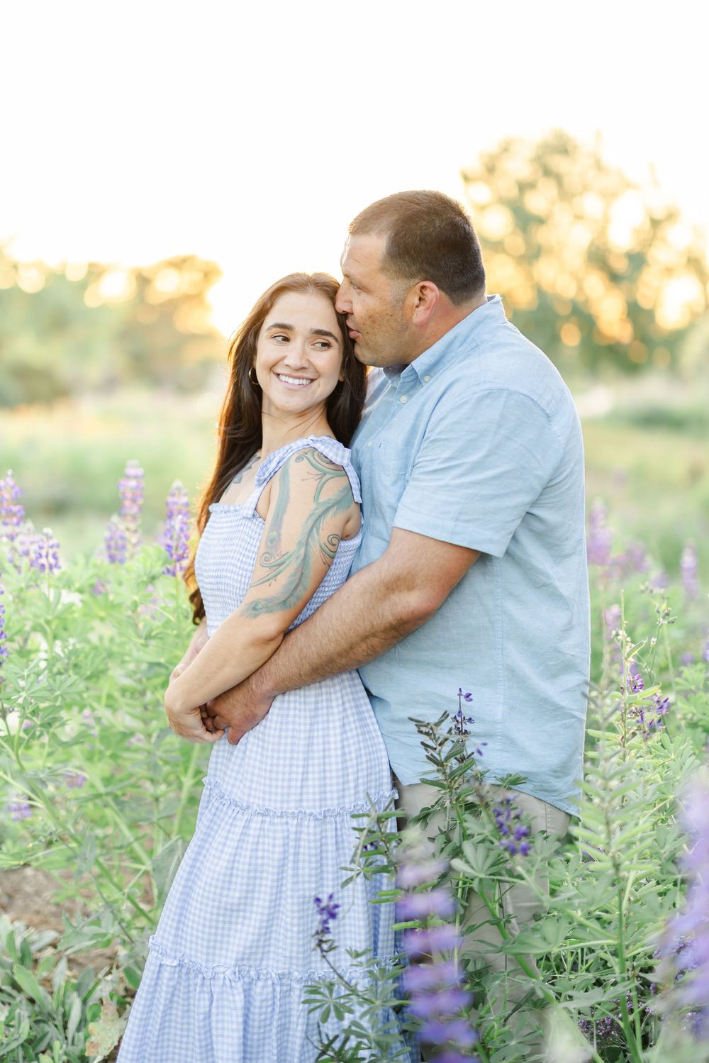 Romantic couple poses among purple lupine flowers in a sun-drenched garden setting.