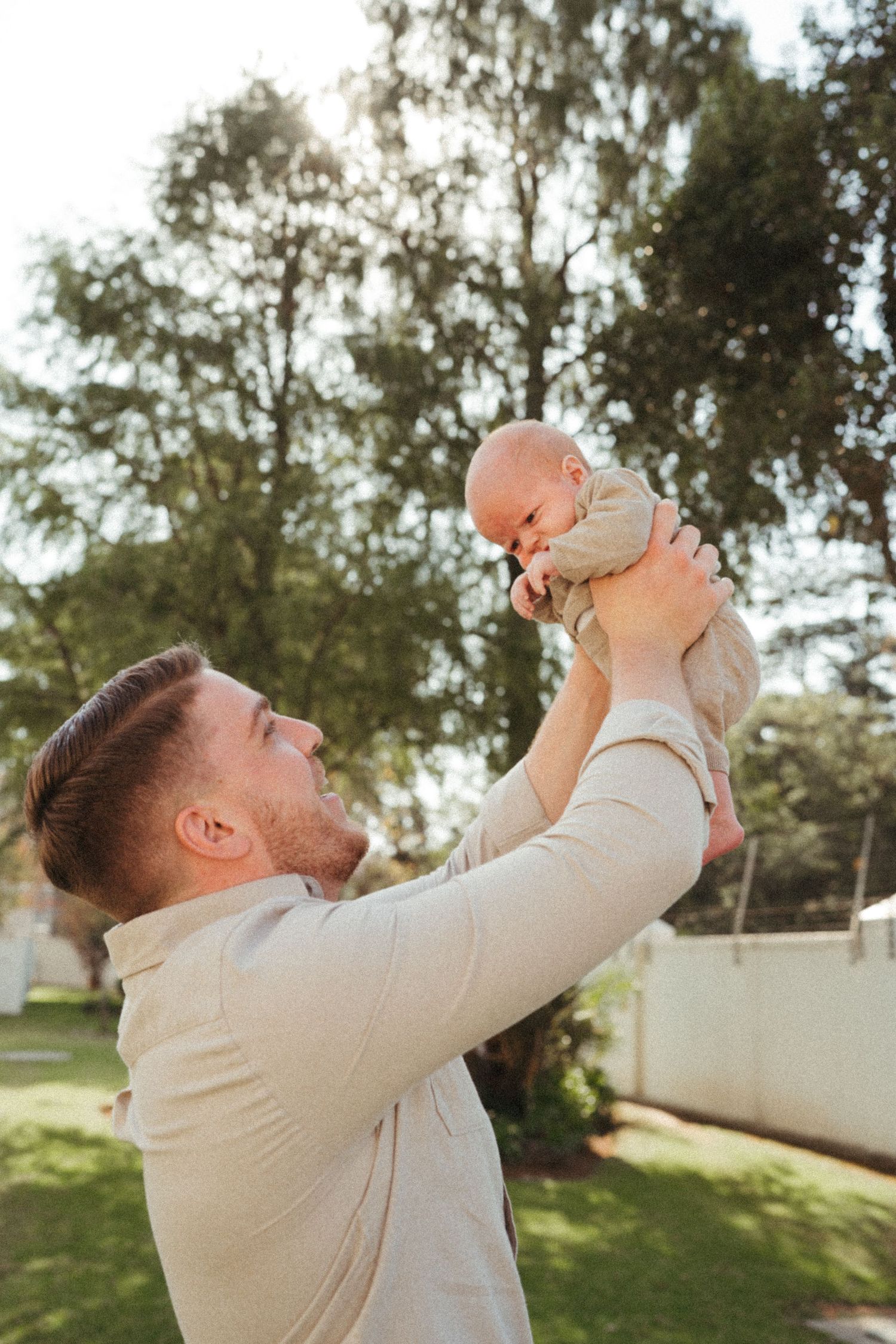 Parent lifting baby joyfully in the air against a natural outdoor backdrop.