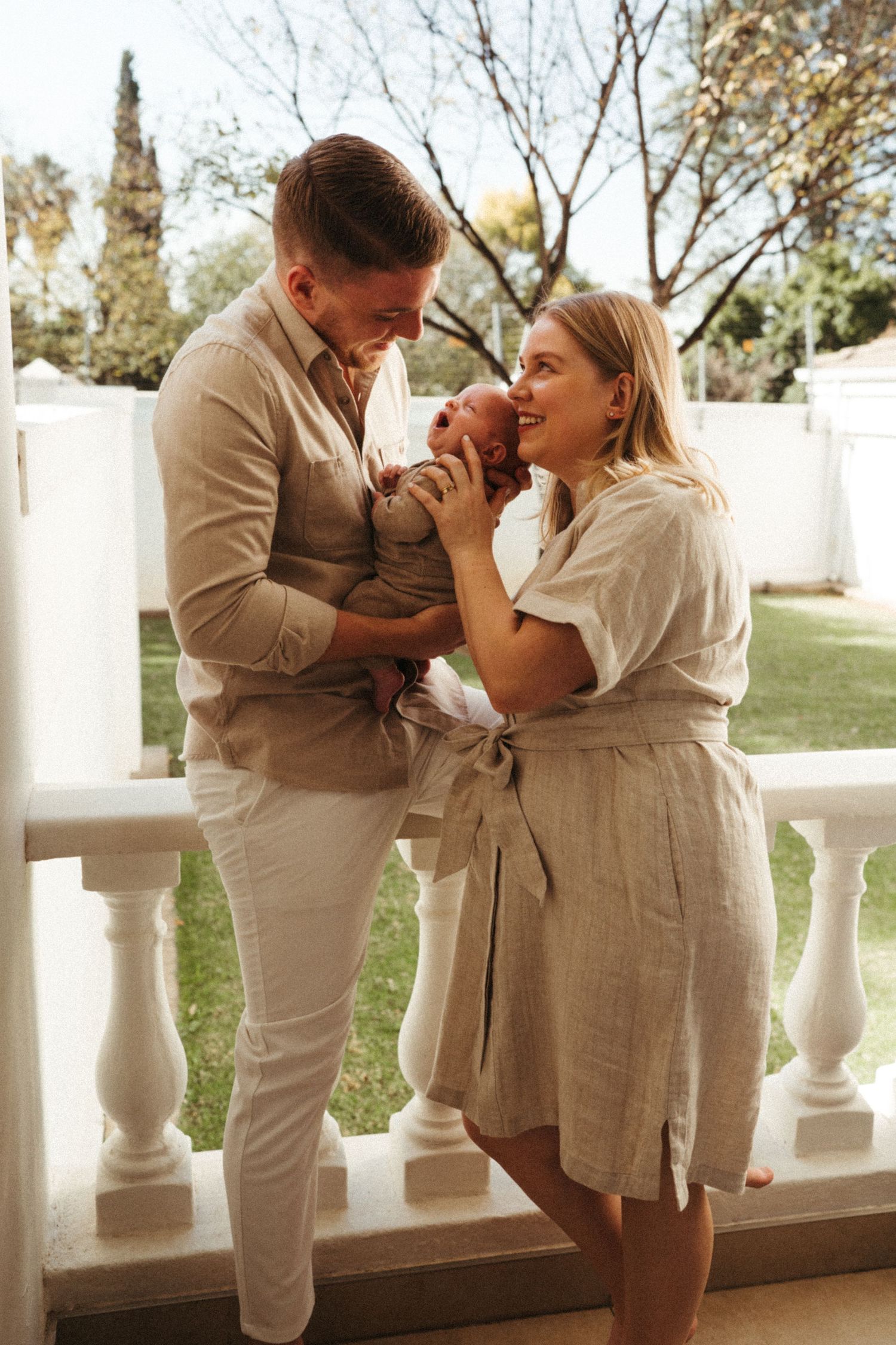 A romantic couple in matching beige outfits share intimate moments on a white balcony with decorative balusters.