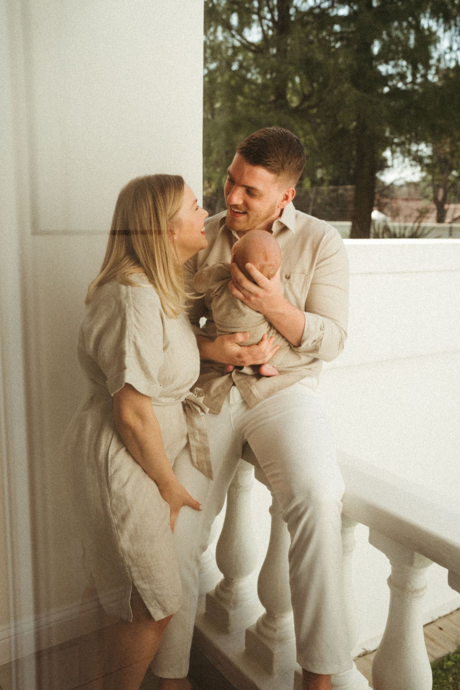A couple in light colored clothing embraces tenderly against a white balcony railing.
