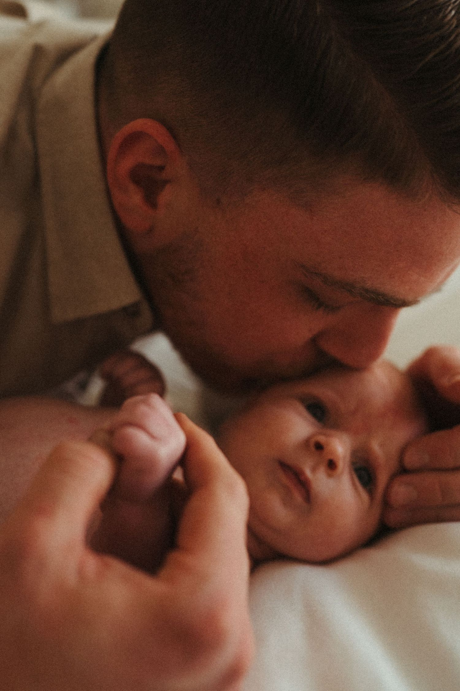 Warm-toned intimate portrait of parent bonding with newborn on white bedding.