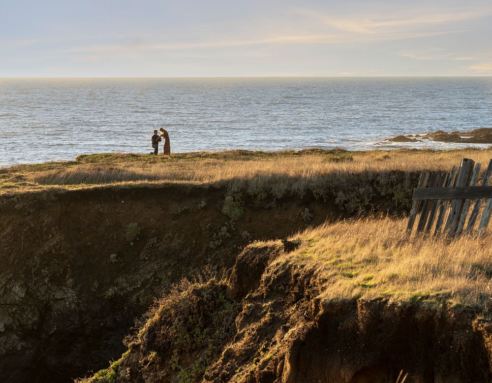 Two figures stand on a dramatic coastal cliff edge at golden hour during surprise engagement.