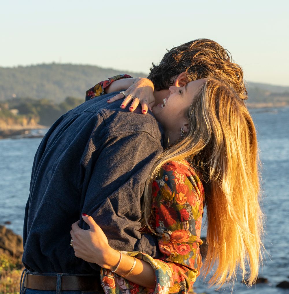 A couple shares an intimate moment on a coastal cliff at sunset during surprise engagement.