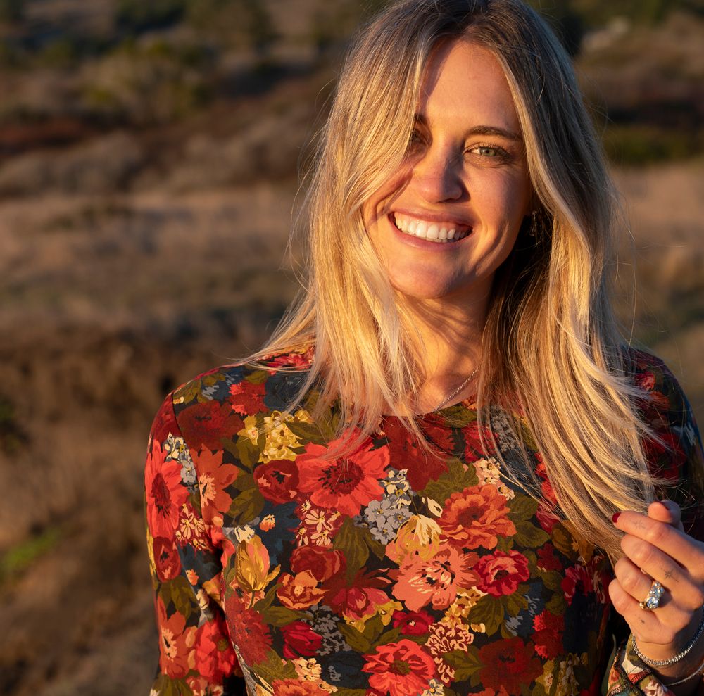 A person with blonde hair wearing a red floral dress poses outdoors during golden hour.