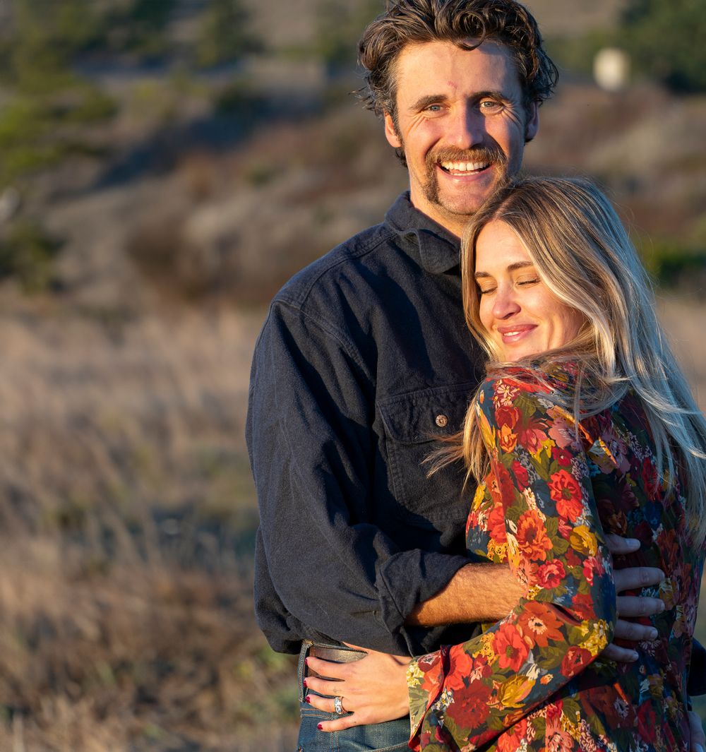 A happy couple embraces during surprise engagement in warm sunset light on a coastal hillside.