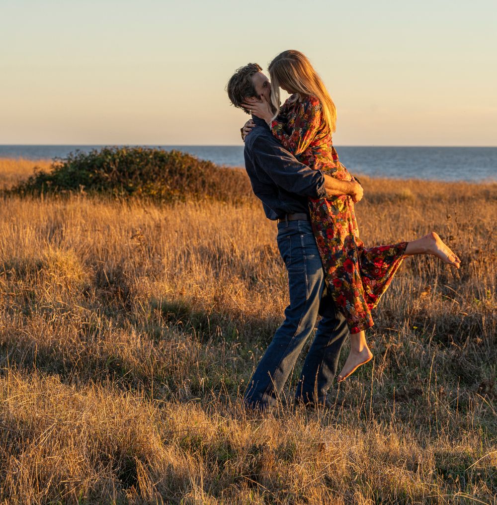 A romantic couple embraces during surprise engagement in golden light on a coastal hillside at sunset.