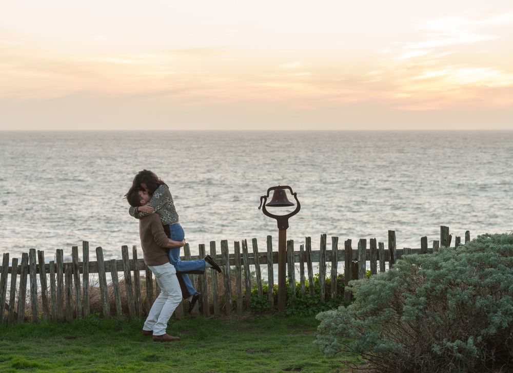 A series of silhouettes against a sunset sky show people watching the ocean from a wooden fence overlook.